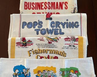 May include: A stack of vintage novelty towels with humorous text and illustrations. The towels include phrases like "Crying Towel Society Member," "Businessman's Crying," "Pop's Crying Towel," "Fisherman's Crying," and "The Tourist's Prayer."