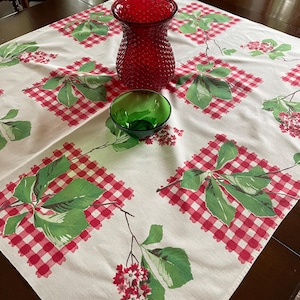 May include: A red hobnail glass vase and a small green bowl sit on a white tablecloth with a red and white gingham pattern. The tablecloth also features green leaves and red flowers.