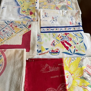 May include: A collection of folded vintage-style cotton kitchen towels featuring colorful screen-printed illustrations. Designs include a map of California, polo players, a person with a donkey, a large yellow sun, and red text reading Pacific Ocean.