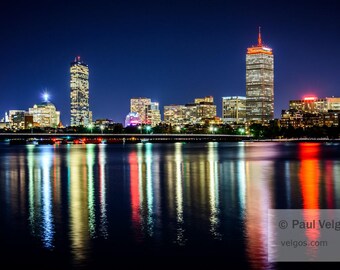 May include: Nighttime cityscape with illuminated skyscrapers reflecting in the water. The buildings have lit windows and are various heights. The sky is dark blue. The water reflects the city lights in shades of orange, yellow, and white.