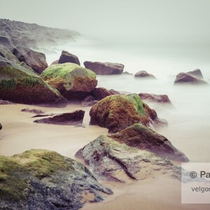 May include: A misty beach scene with large, moss-covered rocks and a sandy shore. The water is blurred and the sky is a light gray.