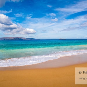 Op de afbeelding: Een schilderachtig strand met goudkleurig zand, turquoise water en een helderblauwe lucht met wolken. De oceaangolven rollen zachtjes op de kust. Een eiland is zichtbaar aan de horizon. De tekst "© Paul Velgos" en "velgos.com" is inbegrepen.