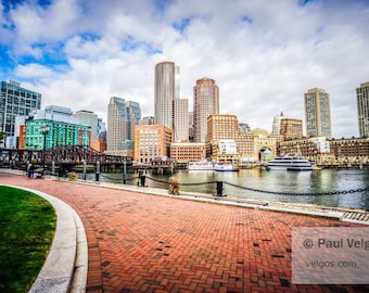 May include: A scenic view of a city skyline with tall buildings, a bridge, and a waterfront. A brick pathway leads towards the water, with boats and a chain fence. The sky is blue with clouds. The image is a landscape photograph.