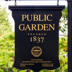 Op de afbeelding: Een zwart bord met gouden letters voor de Public Garden, opgericht in 1837. Het bord bevat het zegel van de stad Boston en de namen van de burgemeester en de commissaris. Het bord hangt aan een zwarte metalen staaf.