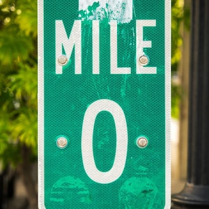 Op de afbeelding: Een groen en wit bord met de tekst "MILE 0" in witte letters. Het rechthoekige bord met afgeronde hoeken is bevestigd aan een metalen paal. De achtergrond is wazig, met groen en een donkere kolom. Het is waarschijnlijk een wegmarkering.