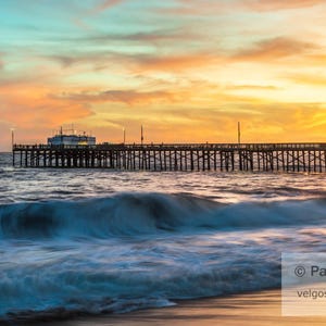 May include: A scenic photograph of a pier extending into the ocean at sunset. The sky is a blend of orange, yellow, and blue hues. Waves crash in the foreground, and the pier's silhouette is visible.