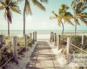Può includere: Una serena scena di spiaggia con una passerella di legno che conduce all'oceano. Le palme incorniciano il sentiero, con sabbia e vegetazione su entrambi i lati. Il cielo è azzurro e l'acqua è turchese chiaro.