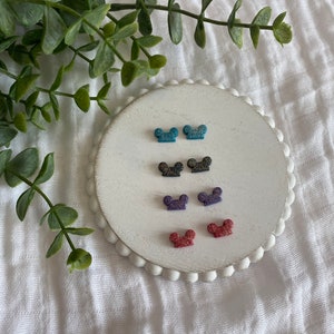 May include: Six pairs of glitter mouse ear stud earrings in a variety of colors, including blue, black, purple, and red. The earrings are displayed on a white circular plate.