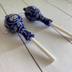 May include: A pair of blue and white lollipop earrings with silver hooks. The lollipops are printed with the words "Tootsie Pop" and "Grape".