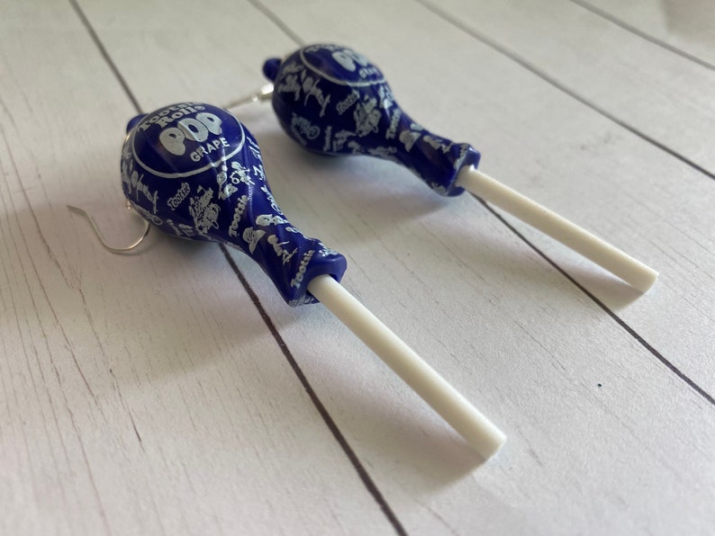 May include: A pair of blue and white lollipop earrings with silver hooks. The lollipops are printed with the words "Tootsie Pop" and "Grape".