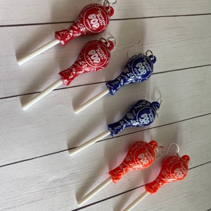 May include: Four pairs of dangle earrings featuring Tootsie Roll Pop lollipops. Two pairs are red with white sticks, one pair is blue with a white stick, and one pair is orange with a white stick.