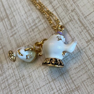 May include: A gold chain necklace with two white porcelain teapot charms. The larger teapot has a purple rim and a gold base. The smaller teapot has a gold spout and handle.