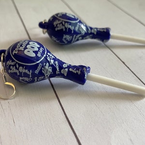 May include: A pair of blue and white earrings shaped like Tootsie Roll Pops. The earrings are made of plastic and have silver hooks.