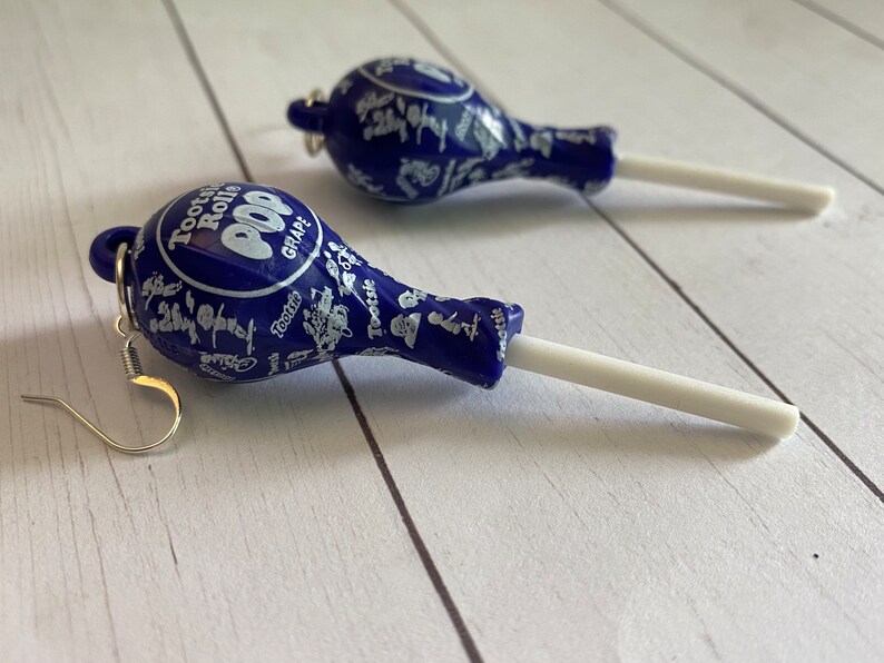May include: A pair of blue and white earrings shaped like Tootsie Roll Pops. The earrings are made of plastic and have silver hooks.