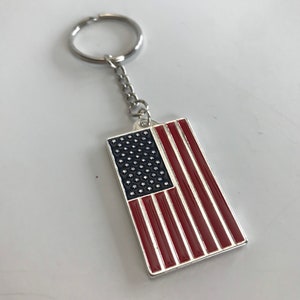 May include: Rectangular keychain featuring the American flag design. The flag has a black field of stars and red and white stripes, with a silver-colored metal border. The keychain includes a chain and a silver-colored ring.