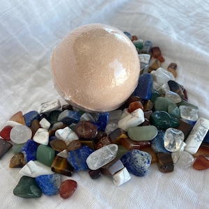 May include: A peach-colored bath bomb wrapped in clear plastic sits atop a bed of colorful polished gemstones. The gemstones are a mix of blues, greens, reds, and whites, creating a vibrant and textured display. The background is a white, textured fabric.