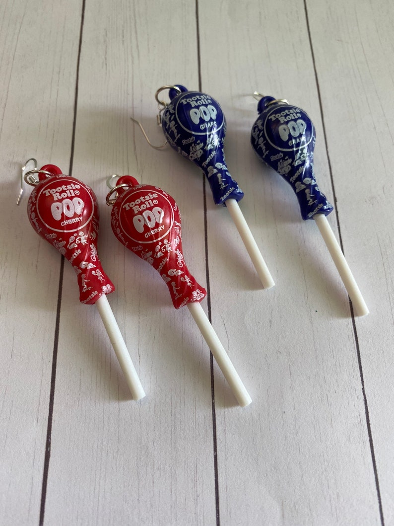 May include: Pair of earrings featuring Tootsie Roll Pop lollipops. One pair is red with cherry flavour and the other is blue with grape flavour.