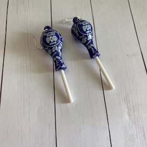 May include: A pair of blue and white Tootsie Roll Pop earrings. The earrings are shaped like lollipops with a white stick and a blue lollipop head. The lollipop head has the Tootsie Roll Pop logo and the flavour "Grape" printed on it.