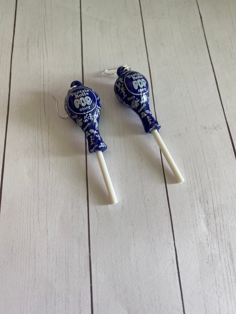 May include: A pair of blue and white Tootsie Roll Pop earrings. The earrings are shaped like lollipops with a white stick and a blue lollipop head. The lollipop head has the Tootsie Roll Pop logo and the flavour "Grape" printed on it.