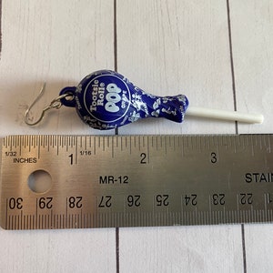 May include: A pair of earrings shaped like a Tootsie Pop lollipop. The lollipop is blue with white and silver details and the stick is white. The earrings are approximately 5 cm long.