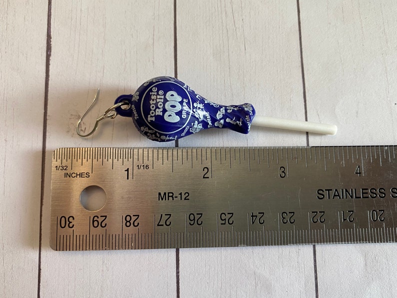 May include: A pair of earrings shaped like a Tootsie Pop lollipop. The lollipop is blue with white and silver details and the stick is white. The earrings are approximately 5 cm long.