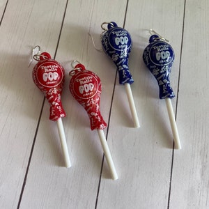 May include: Three pairs of dangle earrings featuring Tootsie Roll Pop lollipops. The lollipops are red and blue with white stick stems. The red lollipops say "Tootsie Roll Pop Cherry" and the blue lollipops say "Tootsie Roll Pop Grape".