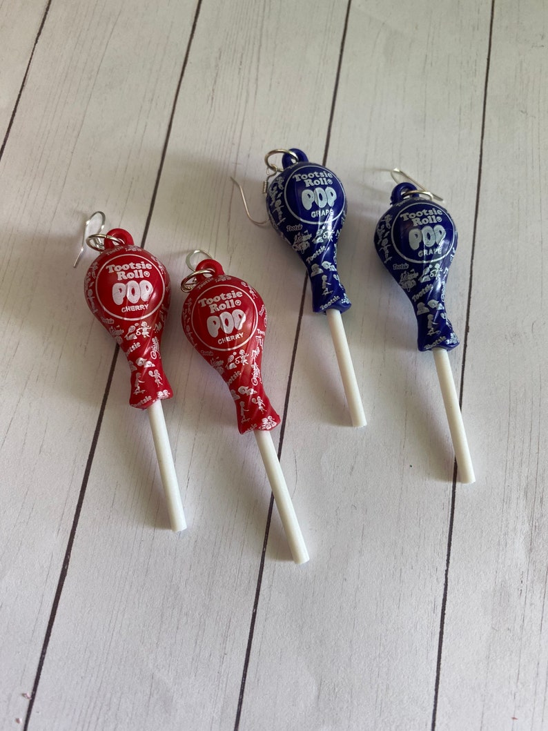 May include: Three pairs of dangle earrings featuring Tootsie Roll Pop lollipops. The lollipops are red and blue with white stick stems. The red lollipops say "Tootsie Roll Pop Cherry" and the blue lollipops say "Tootsie Roll Pop Grape".