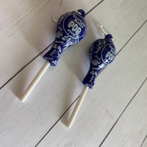 May include: A pair of dangle earrings featuring a blue Tootsie Roll Pop lollipop with a white stick. The lollipop is printed with the words "Tootsie Roll Pop" and "Grape".