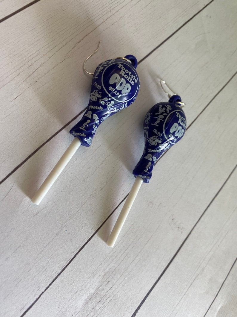 May include: A pair of dangle earrings featuring a blue Tootsie Roll Pop lollipop with a white stick. The lollipop is printed with the words "Tootsie Roll Pop" and "Grape".