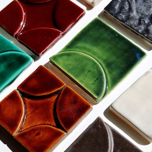 May include: A selection of square ceramic tiles in shades of red, green, brown, and white. Some tiles have a curved pattern within the square. The tiles are displayed on a white surface, highlighting their glossy finish and varied textures.