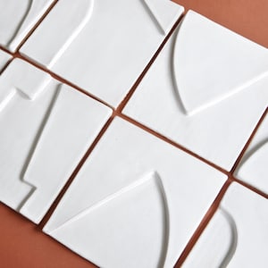Op de afbeelding: Een set van negen witte keramische tegels met geometrische reliëfmotieven. Elke tegel heeft een ander abstract ontwerp, wat een moderne en minimalistische esthetiek creëert.