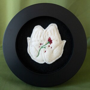 May include: Two white felt hands holding a single red flower with a green stem. The hands and flower are embroidered on a black background and framed in a black circular frame.
