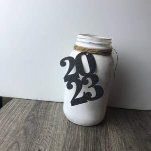 Puede incluir: Tarro de alba&ntilde;il blanco pintado con un recorte de 2023 de purpurina negra sujeto con un cordel.