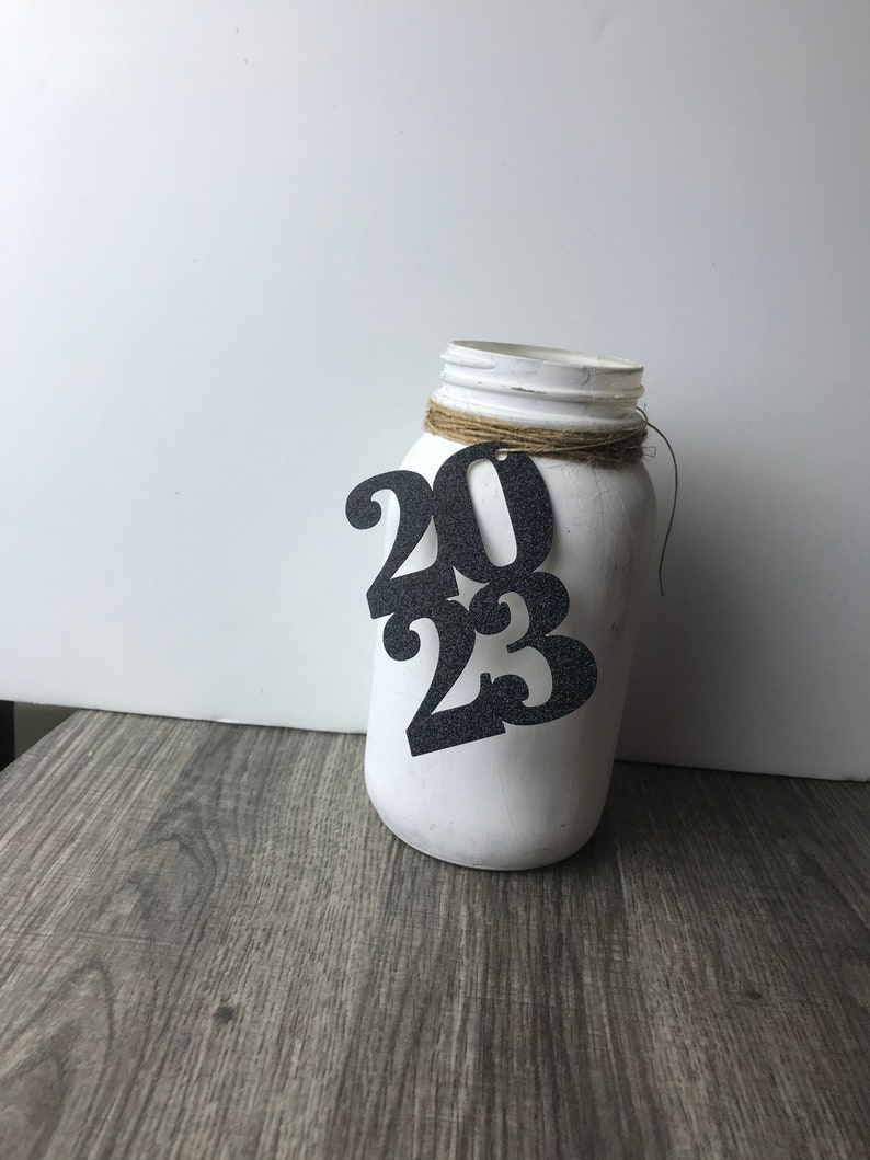 Puede incluir: Tarro de alba&ntilde;il blanco pintado con un recorte de 2023 de purpurina negra sujeto con un cordel.
