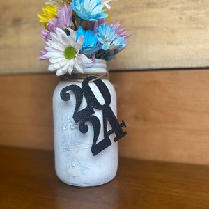 Puede incluir: Un tarro de alba&ntilde;il blanco pintado con un lazo de arpillera y un recorte de papel negro del n&uacute;mero 2024. El tarro est&aacute; lleno de un ramo de flores amarillas, azules y rosas.
