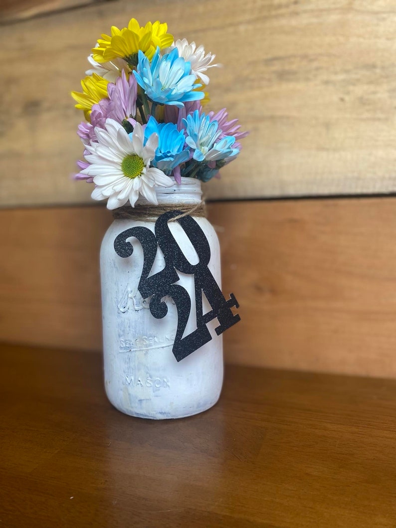 Puede incluir: Un tarro de alba&ntilde;il blanco pintado con un lazo de arpillera y un recorte de papel negro del n&uacute;mero 2024. El tarro est&aacute; lleno de un ramo de flores amarillas, azules y rosas.