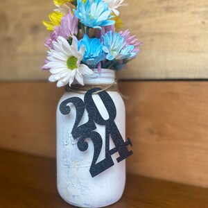 Puede incluir: Un tarro de alba&ntilde;il blanco pintado con un letrero de 2024 de purpurina negra, lleno de un ramo de flores amarillas, blancas, azules y moradas.