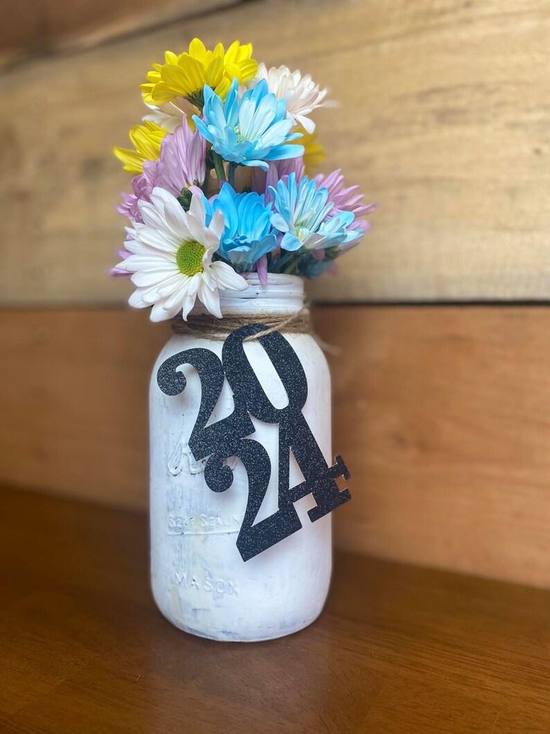 Puede incluir: Un tarro de alba&ntilde;il blanco pintado con un letrero de 2024 de purpurina negra, lleno de un ramo de flores amarillas, blancas, azules y moradas.