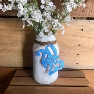 Puede incluir: Un tarro de cristal blanco con una decoraci&oacute;n de purpurina azul 2023, lleno de flores de aliento de beb&eacute; blancas.