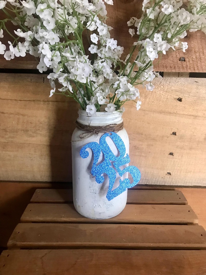 Puede incluir: Un tarro de cristal blanco con una decoraci&oacute;n de purpurina azul 2023, lleno de flores de aliento de beb&eacute; blancas.
