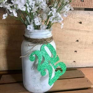 Puede incluir: Tarro de alba&ntilde;il blanco con un letrero de purpurina verde 2023 pegado a &eacute;l. El tarro est&aacute; lleno de flores blancas.