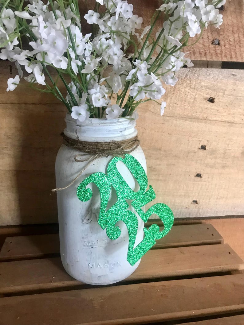 Puede incluir: Tarro de alba&ntilde;il blanco con un letrero de purpurina verde 2023 pegado a &eacute;l. El tarro est&aacute; lleno de flores blancas.
