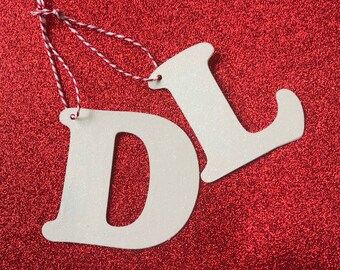 May include: Two white glitter letters, "D" and "L", are strung together with red and white twine on a red glitter background.