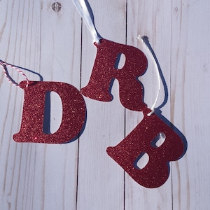 May include: Three red glitter letters, D, R, and B, are strung together with white twine. Each letter has a white ribbon attached to the top.