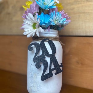Puede incluir: Un tarro de cristal blanco con una cinta de arpillera y un letrero de purpurina negra que dice 2024. El tarro est&aacute; lleno de un ramo de flores amarillas, blancas, azules y moradas.
