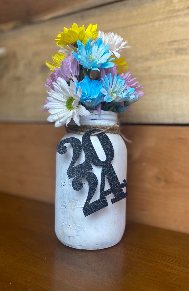 Puede incluir: Un tarro de cristal blanco con una cinta de arpillera y un letrero de purpurina negra que dice 2024. El tarro est&aacute; lleno de un ramo de flores amarillas, blancas, azules y moradas.