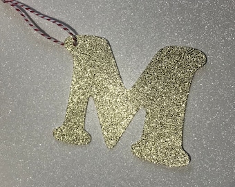 May include: Gold glitter letter M ornament with a red and white string for hanging.
