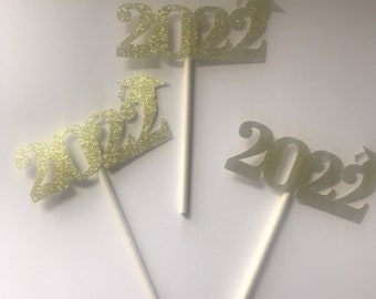 May include: Three gold glitter 2022 graduation cake toppers with a graduation cap on the 2. Each topper is attached to a white plastic stick.