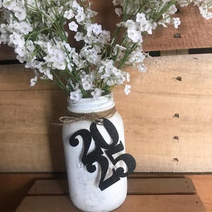 Puede incluir: Un tarro de alba&ntilde;il blanco pintado con una cinta de arpillera y un letrero de 2023 con purpurina negra. El tarro est&aacute; lleno de flores blancas de aliento de beb&eacute;.