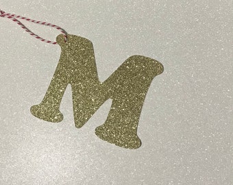 May include: Gold glitter letter M ornament with a red and white twine string.
